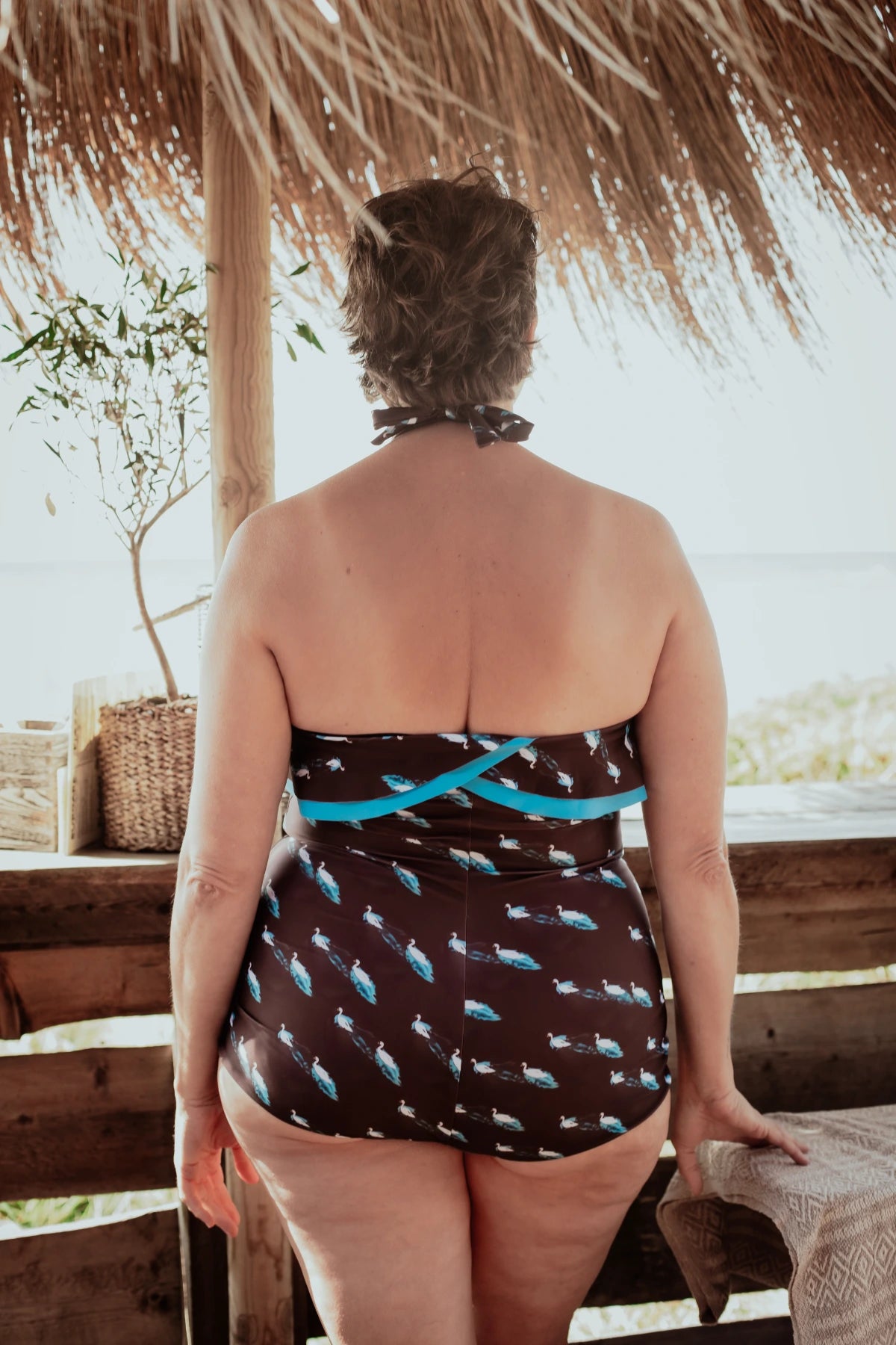 Woman wearing a swans patterned swimsuit with ruffles. Standing under a thatched roof.