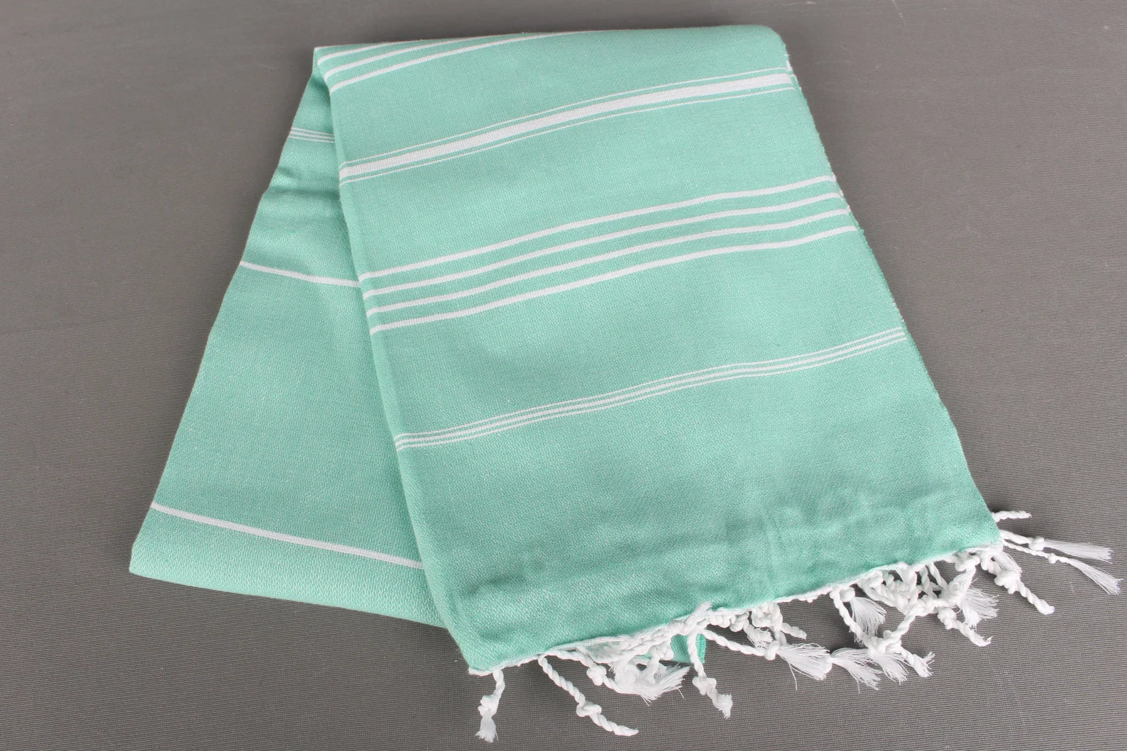 Turquoise lightweight hamam towel with white stripes and fringes on a gray background