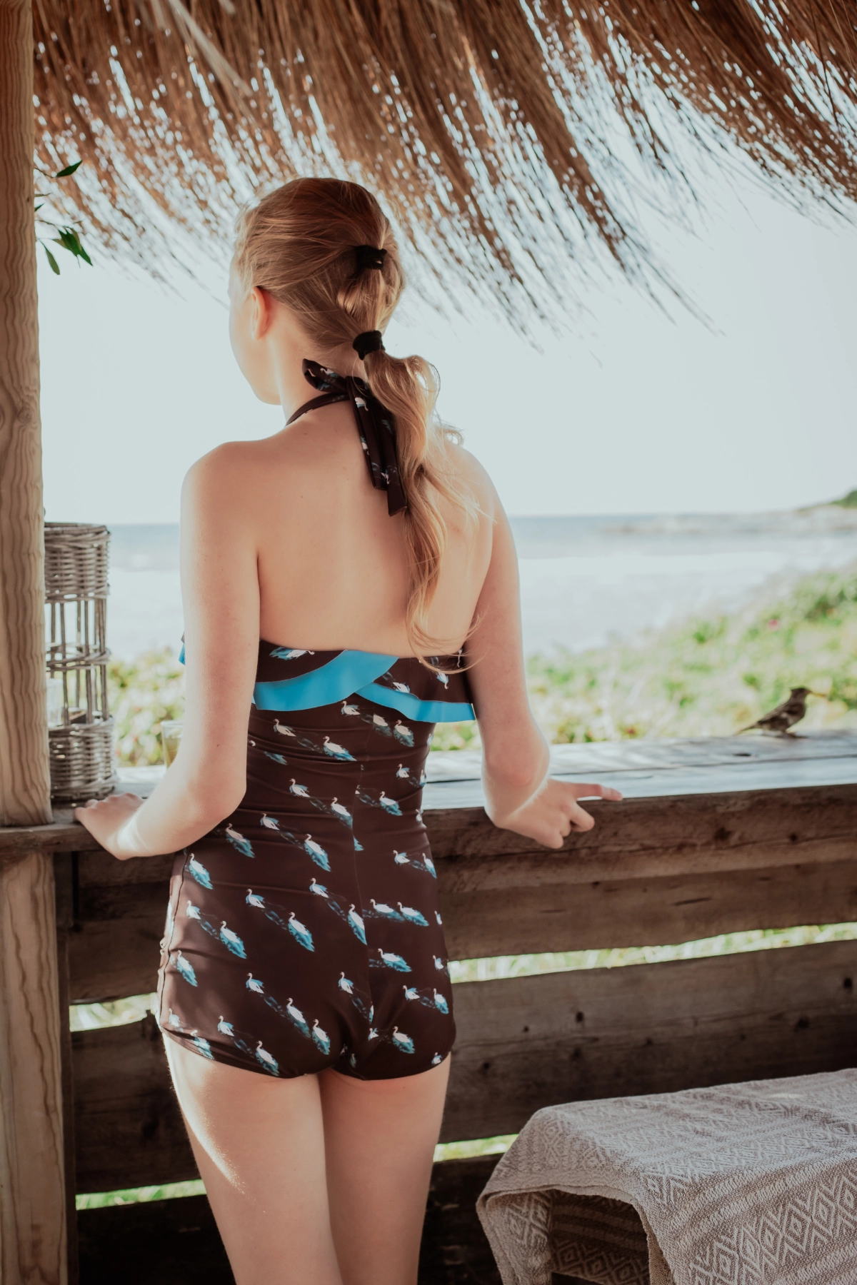 Young woman in Dancer ruffled swimsuit in swans print. Standing by a wooden structure with a scenic view and little Disney looking bird to the right.