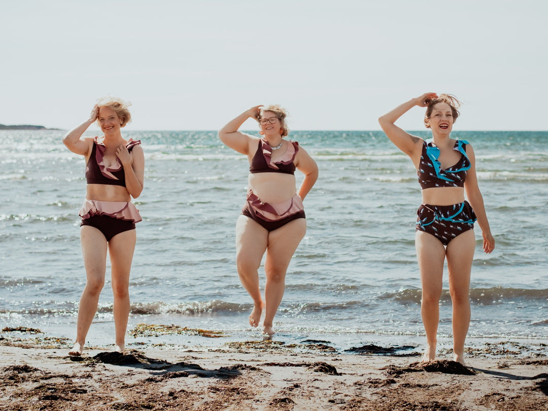 Three women in ruffled bikinis standing on a beach in heavy wind.  Ocean in the background