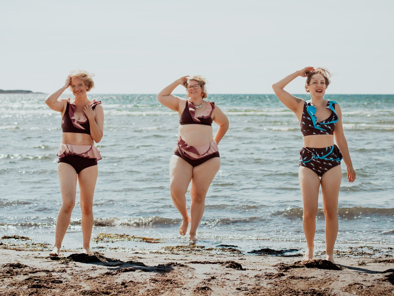 Three women in ruffled bikinis standing on a beach in heavy wind.  Ocean in the background